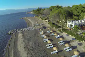 Campingplatz am See mit Kiesstrand, Sonnenliegen und Sonnenschirmen, Bäumen und einem Gebäude am Ufer.