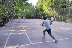 Tennisplatz auf dem Campinggelände mit Netz, Linien und Spielern im Hintergrund, umgeben von Bäumen und Hecken.