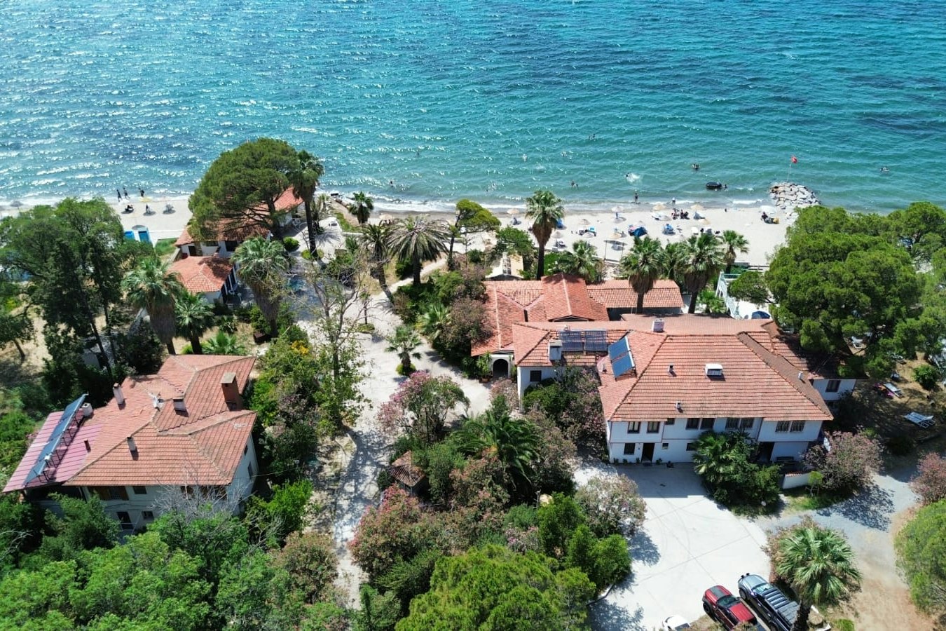 Luftaufnahme eines Campingplatzes am Meer mit mehreren Gebäuden, roten Dächern, Wegen, Bäumen und einem Parkplatz mit Auto.