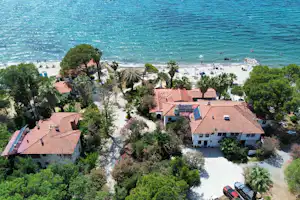 Luftaufnahme eines Campingplatzes am Meer mit mehreren Gebäuden, roten Dächern, Wegen, Bäumen und einem Parkplatz mit Auto.