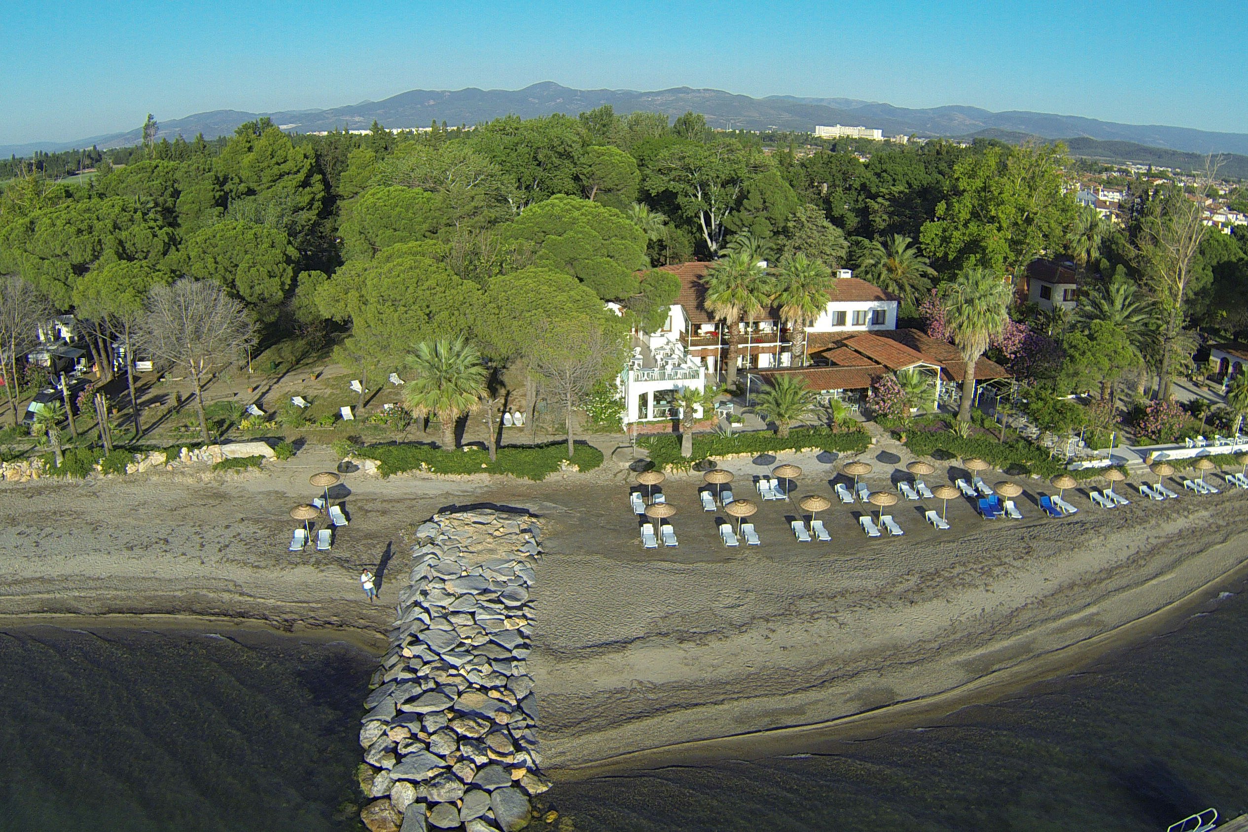 Luftaufnahme eines Campingplatzes am Strand mit Reihen von Liegestühlen, Steinterrassen und einem Gebäude im Grünen.