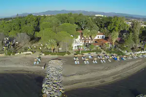 Luftaufnahme eines Campingplatzes am Strand mit Reihen von Liegestühlen, Steinterrassen und einem Gebäude im Grünen.