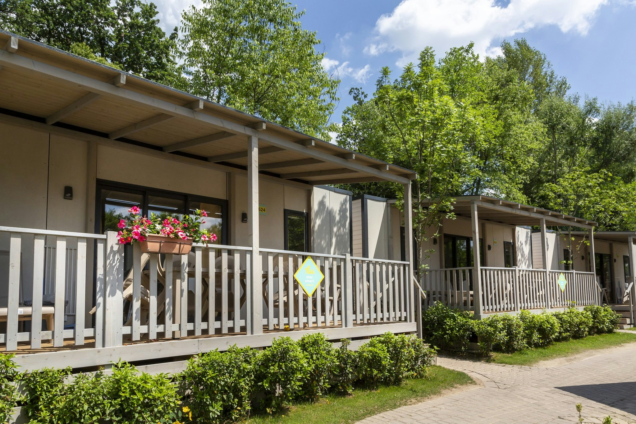 Reihen von Ferienhäusern mit Holzgeländer und Veranda, Blumenampel und gepflegtem Gartenweg, umgeben von grünen Bäumen.