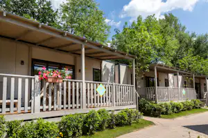 Reihen von Ferienhäusern mit Holzgeländer und Veranda, Blumenampel und gepflegtem Gartenweg, umgeben von grünen Bäumen.