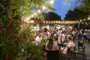Gelateria am Campingplatz mit beleuchteten Lichtketten, Gästen an Tischen und Stühlen vor dem Verkaufsstand.
