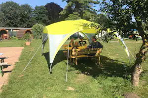 Gelbes Zelt auf dem Campingplatz mit Holztisch und Sitzbank, umgeben von Rasen, Bäumen und weiteren Unterkünften.