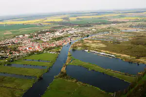 Luftaufnahme eines Flusstals mit Dorf, grünen Wiesen und einem Kanal mit Stegen und Booten.