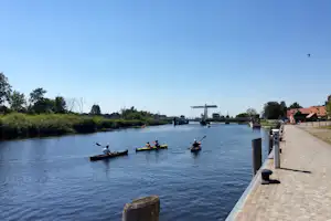 Kajaks auf dem Kanal, Paddelboote im Wasser, Steg und Uferweg mit Bäumen und Häusern im Hintergrund.