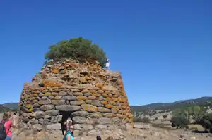 Steinmauer mit Öffnung und Baumkrone oben, im Hintergrund Hügel und blauer Himmel.