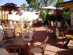 Terrasse mit vielen Holzstühlen und Tischen unter Sonnenschirm, im Hintergrund Blumenampeln und ein Gebäude am Campingplatz.