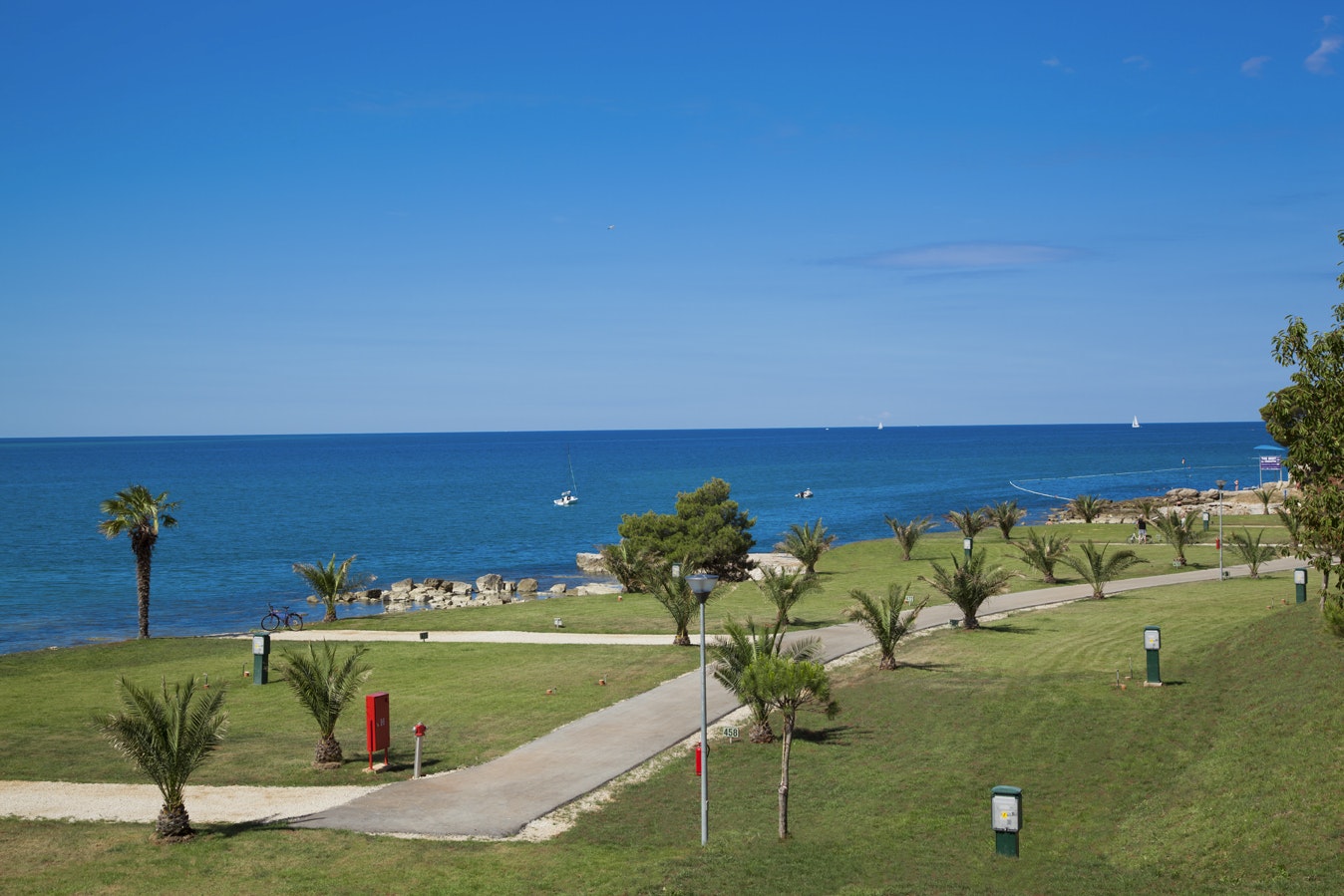 Blick auf den Strand und das Meer von einer gepflegten Campinganlage mit Rasen, Wegen und kleinen Bäumen.