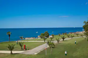 Blick auf den Strand und das Meer von einer gepflegten Campinganlage mit Rasen, Wegen und kleinen Bäumen.