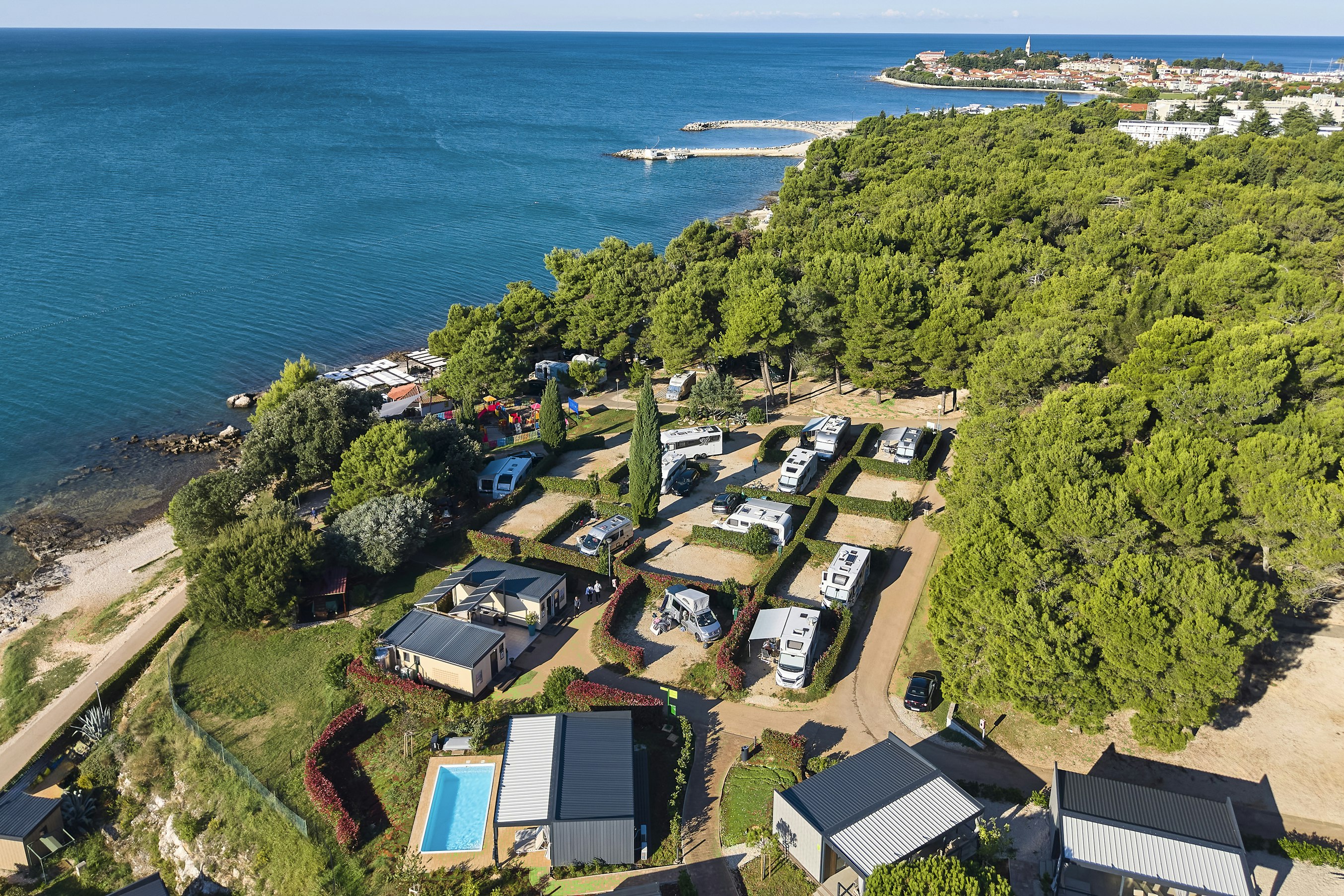 Luftaufnahme eines Campingplatzes am Meer mit Stellplätzen, kleinen Gebäuden, Pool und Küstenlinie mit Bäumen.