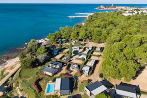 Luftaufnahme eines Campingplatzes am Meer mit Stellplätzen, kleinen Gebäuden, Pool und Küstenlinie mit Bäumen.
