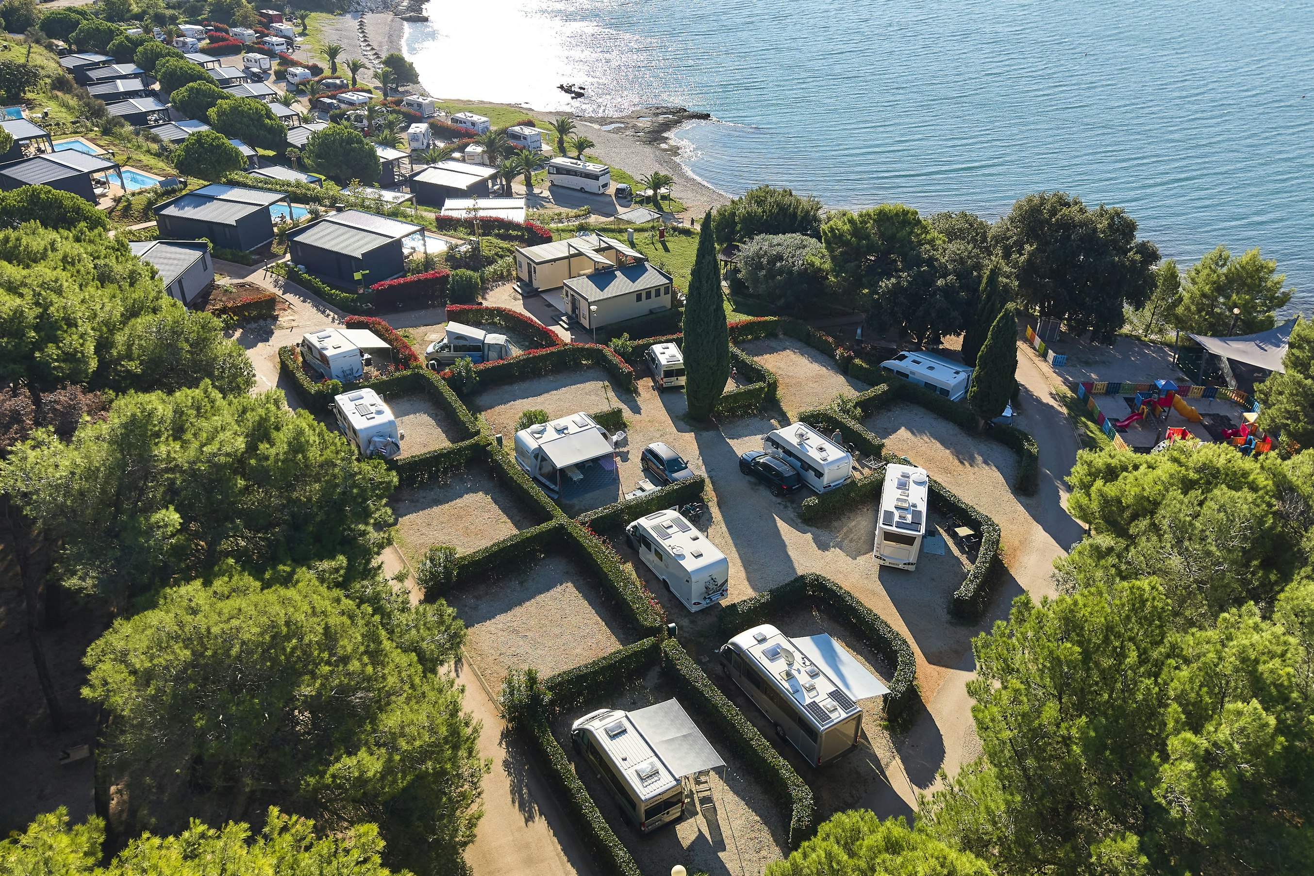 Luftaufnahme eines Campingplatzes mit Wohnmobilen und Stellplätzen, umgeben von Bäumen, nahe dem Meer und einer Bucht.