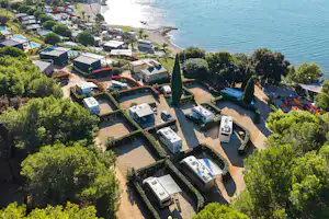 Luftaufnahme eines Campingplatzes mit Wohnmobilen und Stellplätzen, umgeben von Bäumen, nahe dem Meer und einer Bucht.