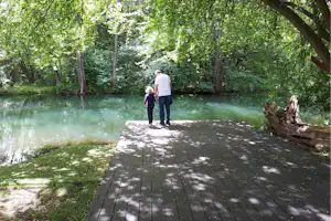 Holzsteg am See im Grünen, zwei Personen stehen am Ufer, im Hintergrund Bäume und Wasser.