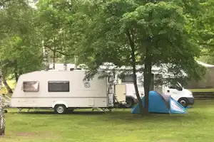 Wohnwagen und Zelt auf einem Campingplatz, umgeben von Bäumen, mit Rasenfläche und geparktem Auto im Hintergrund.