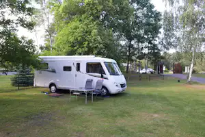 Weißes Wohnmobil auf einem Campingplatz im Grünen, mit Vorzelt und Tisch am Stellplatz, umgeben von Bäumen.