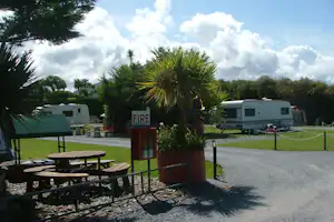 Campingplatz mit Wohnwagen, Kiesweg und Feuerlöscher-Schild, im Hintergrund Palmen und Bäume.