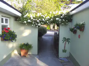Gepflasterter Weg zwischen weißen Hauswänden mit Blumenkästen, hängenden Blumentöpfen und einem Durchgang zum Garten.
