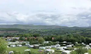 Campingplatz im Tal mit Wohnwagen und Stellplätzen, umgeben von grünen Wiesen, Bäumen und Hügeln in der Ferne.