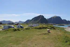 Campingplatz mit Wohnwagen auf einer Wiese, im Hintergrund Berge und ein Seeufer.
