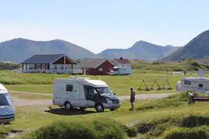 Wohnmobil auf einem Campingplatz mit Rasenfläche, daneben weitere Fahrzeuge und Hütten, im Hintergrund Berge.