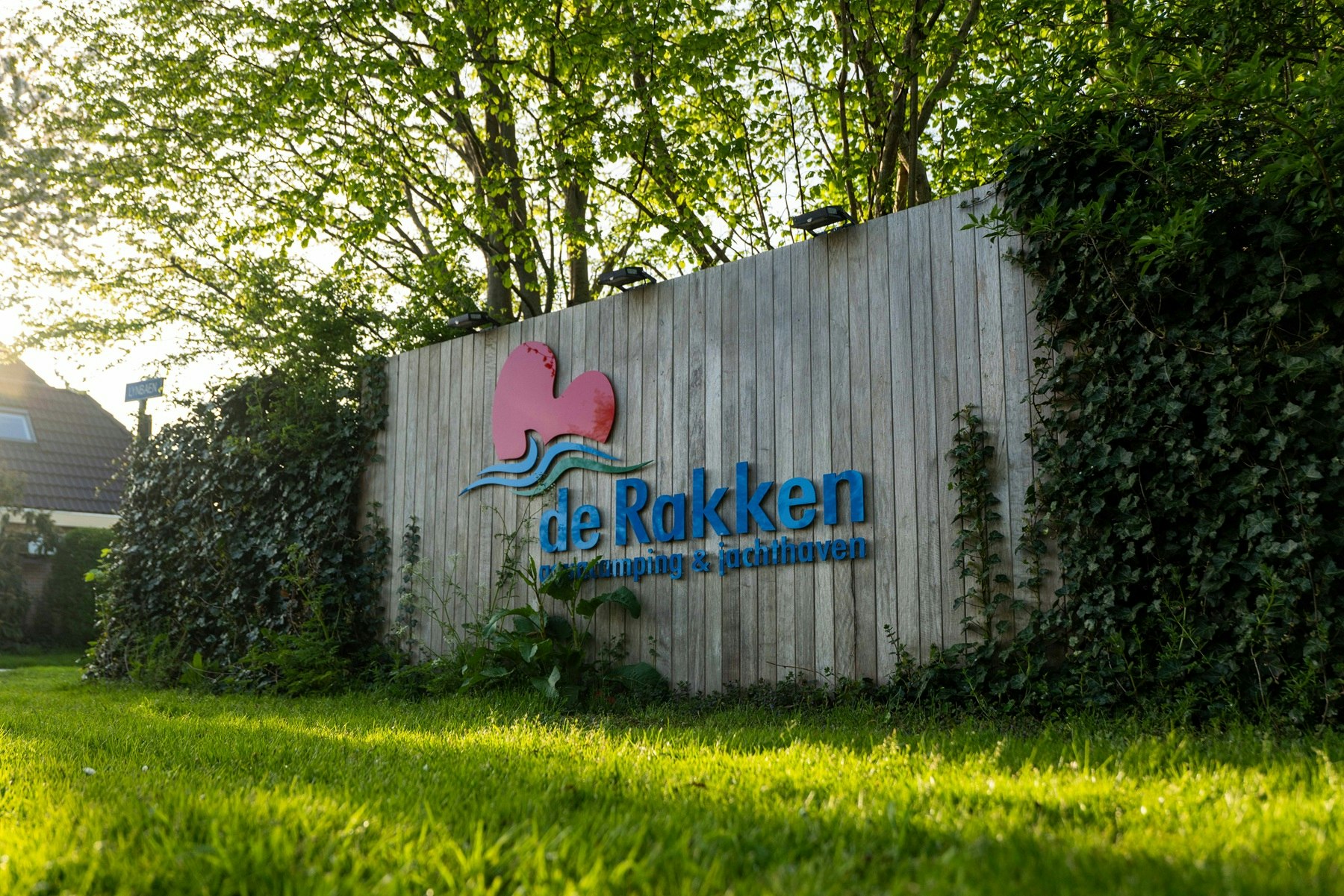 Holzwand mit dem Schriftzug „de Rakken“ und Logo, umgeben von Hecke und Rasen im Campingbereich.
