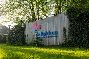 Holzwand mit dem Schriftzug „de Rakken“ und Logo, umgeben von Hecke und Rasen im Campingbereich.