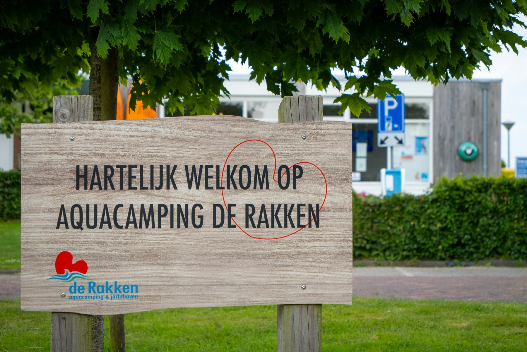 Schild am Campingplatz mit Text „Hartelijk welkom op Aquacamping de Rakken“ vor Hecke und Weg, im Hintergrund ein Parkplatzschild.