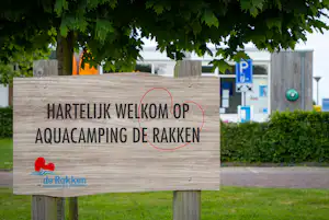 Schild am Campingplatz mit Text „Hartelijk welkom op Aquacamping de Rakken“ vor Hecke und Weg, im Hintergrund ein Parkplatzschild.