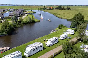 Wohnmobile auf einem Campingplatz am Fluss, mit Stellplätzen, Bäumen und Häusern am Ufer, Blick von oben.