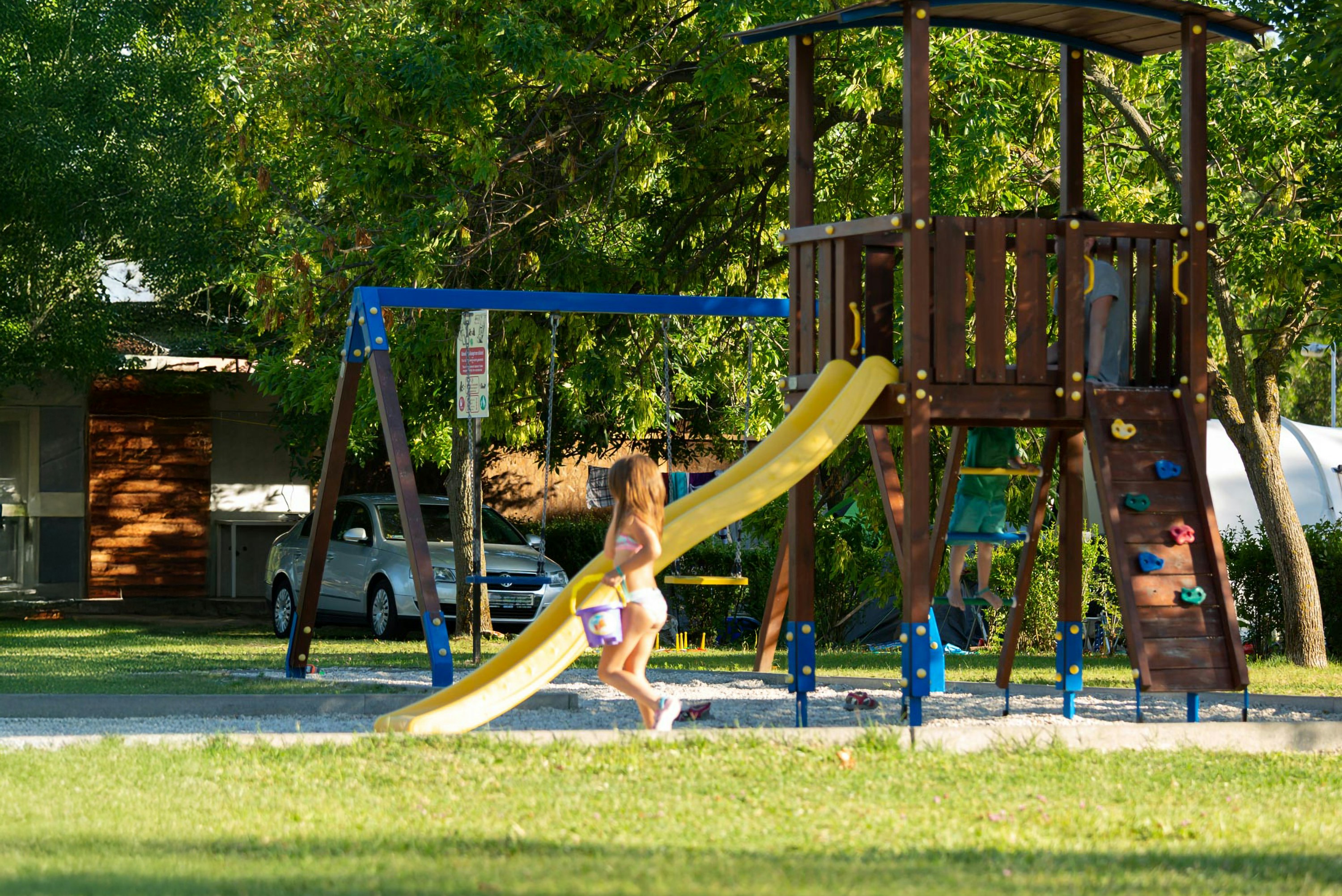 Spielplatz im Campingpark mit Rutsche und Klettergerüst, Kind läuft über den Rasen, im Hintergrund Autos und Bäume.