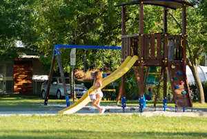 Spielplatz im Campingpark mit Rutsche und Klettergerüst, Kind läuft über den Rasen, im Hintergrund Autos und Bäume.
