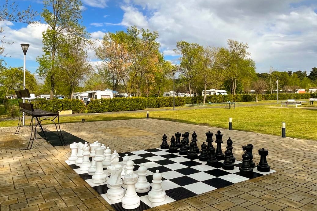 Schachspiel auf einem gepflasterten Terrassenbereich, im Hintergrund Rasen, Bäume und Wohnwagen auf dem Campingplatz.