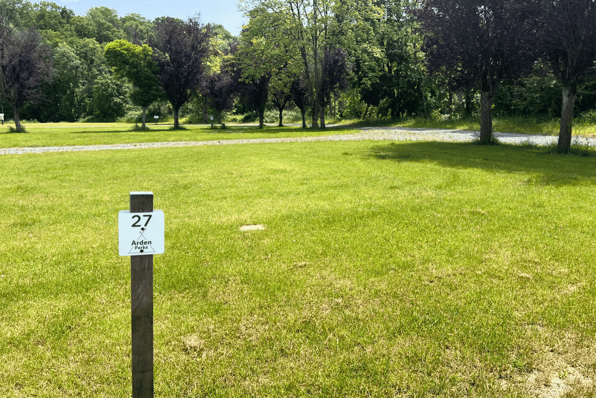 Grüner Campingplatz mit gepflegtem Rasen, Baumreihe im Hintergrund und Stellplatznummer 27 auf einem Schild.