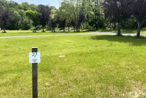 Grüner Campingplatz mit gepflegtem Rasen, Baumreihe im Hintergrund und Stellplatznummer 27 auf einem Schild.
