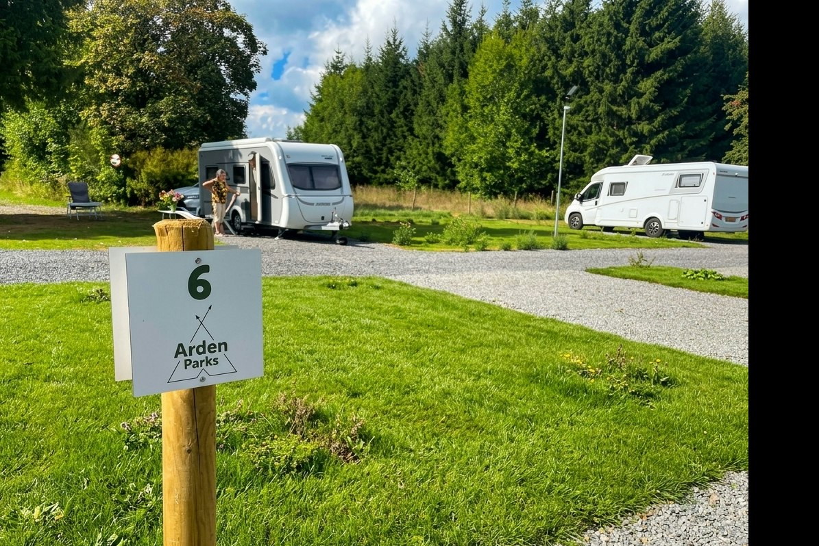 Wohnmobile auf einem Campingplatz, im Vordergrund ein Schild „Arden Parks“ an einem Holzpfosten, daneben Gras und Kiesweg.