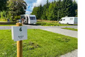 Wohnmobile auf einem Campingplatz, im Vordergrund ein Schild „Arden Parks“ an einem Holzpfosten, daneben Gras und Kiesweg.