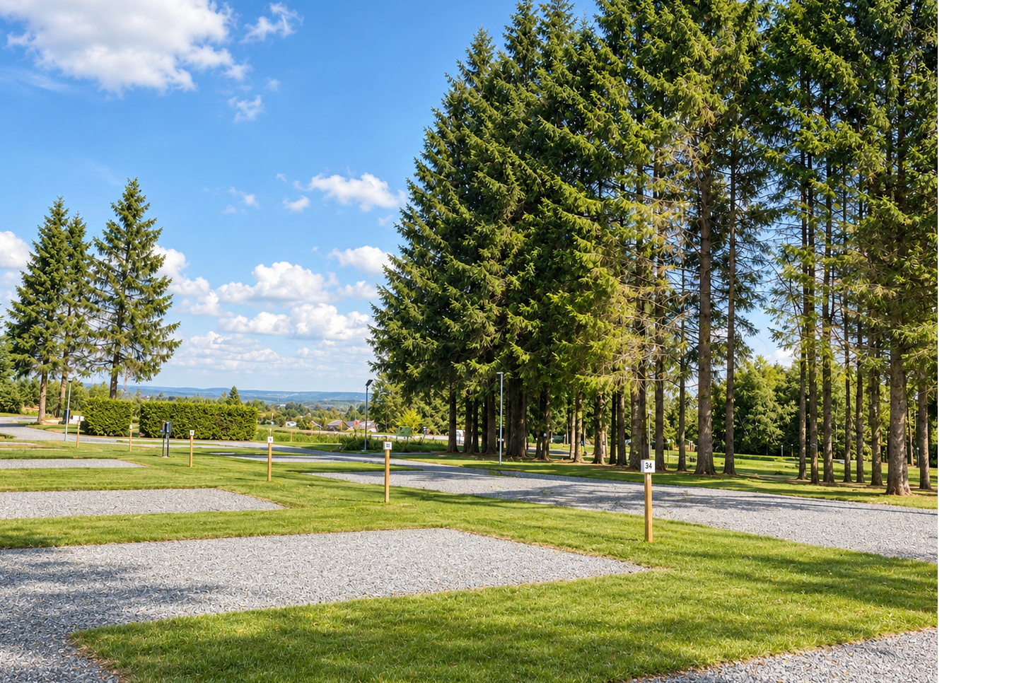 Grüner Campingplatz mit gepflegten Rasenflächen, Kieswegen und hohen Nadelbäumen vor weitem Blick in die Landschaft.