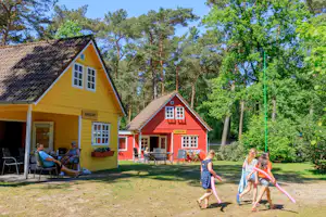 Gelbes und rotes Ferienhaus mit Terrasse, Gartenmöbeln und Schildern, im Grünen zwischen Bäumen auf dem Campingplatz.