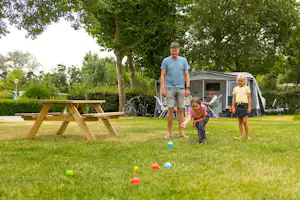 Familie auf dem Campingplatz: Mann und Kinder laufen auf der Wiese vor einem Zelt mit Fahrrädern und Picknickbank.