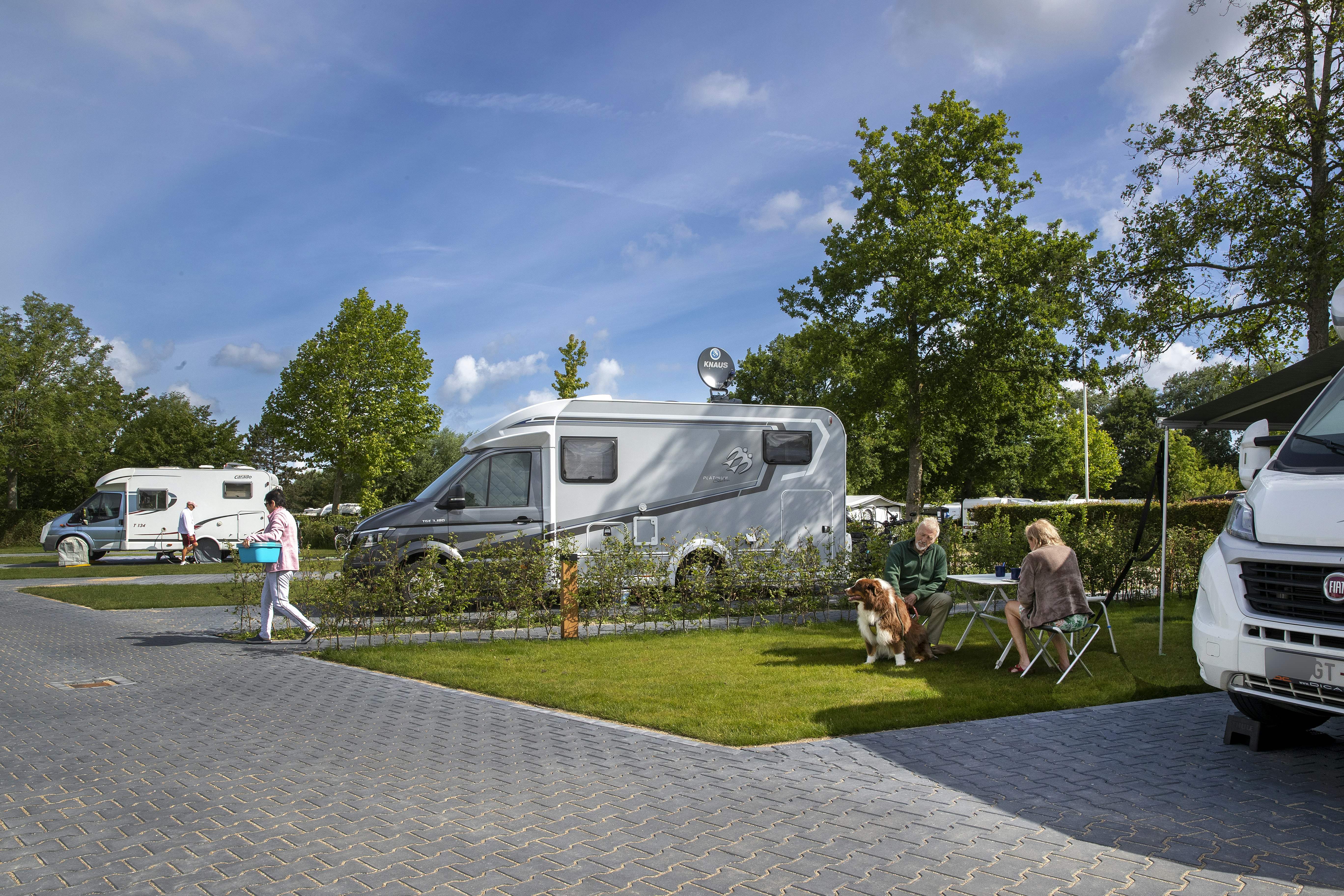 Wohnmobil auf dem Campingplatz-Stellplatz, daneben Wohnwagen und Sitzgruppe auf dem Rasen, Bäume und Wege im Hintergrund.