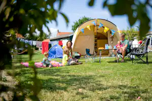Familie im gelben Zelt auf dem Campingplatz, daneben Campingstühle und Spielzeug auf der Wiese vor dem Wohnwagen.