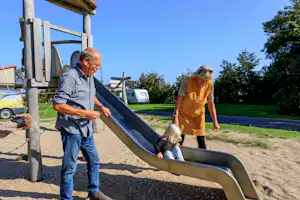 Familie am Campingplatz: Kind rutscht die Rutsche, Erwachsene stehen daneben auf dem Spielbereich.
