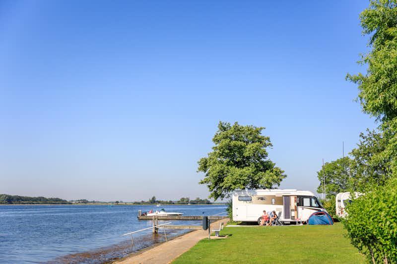 Ardoer Vakantiepark De Paardekreek -  Sitzender Camper vor dem Wohnmobil auf der Wiese der Campingplatzanlage mit Blick aufs Wasser 