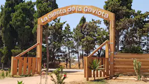 Holzbogen mit Aufschrift „La siesta de la gravita“ über einem Weg, umgeben von Bäumen und Pflanzen im Campingbereich.