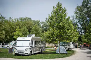 Wohnmobil und Zelt auf einem Campingplatz, daneben weitere Fahrzeuge, im Hintergrund Bäume und ein Weg.