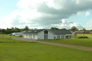 Campinggebäude mit großen Fenstern, Rasenfläche und Schotterweg, im Hintergrund weitere Wohnwagen und Häuser.